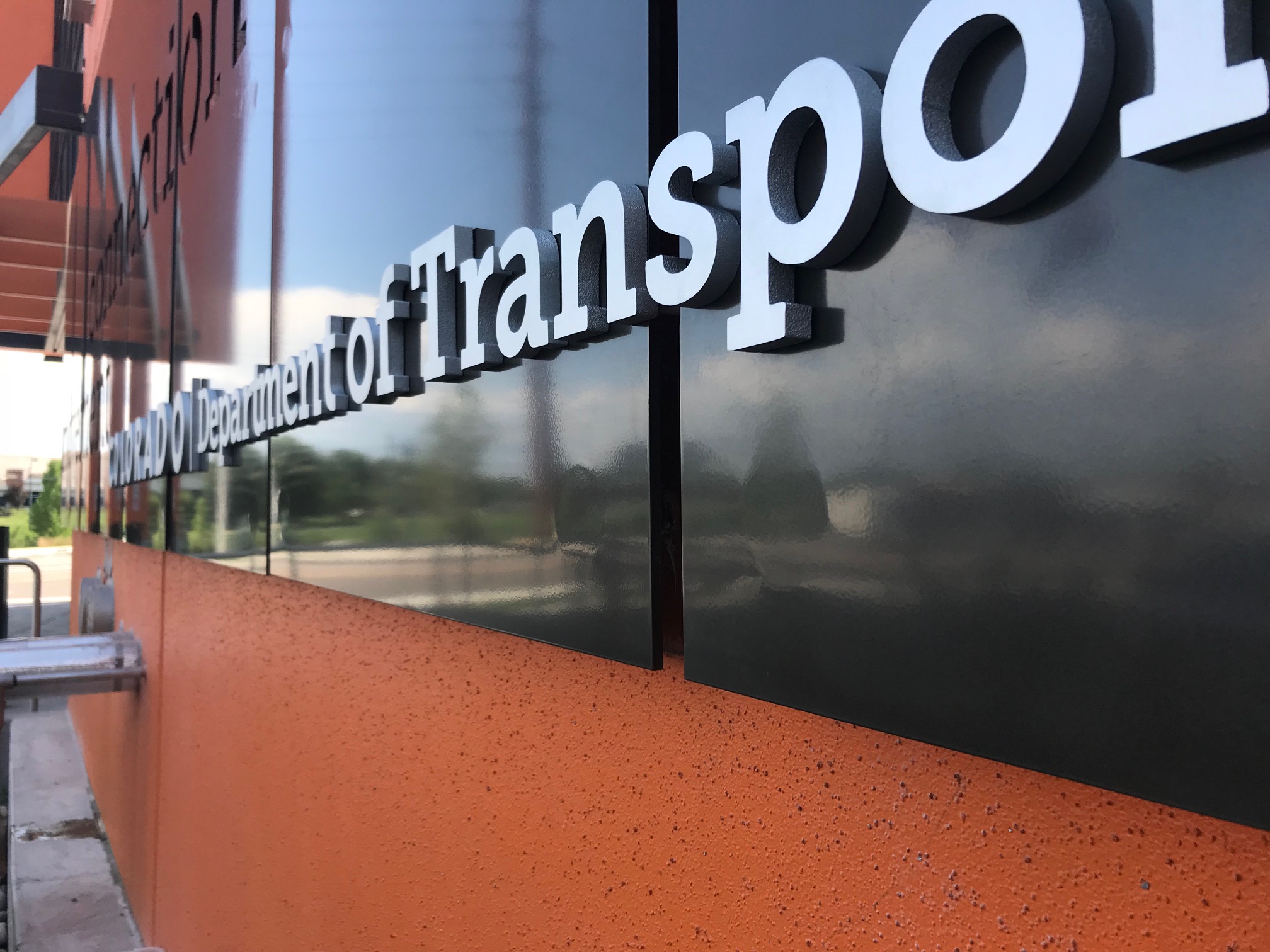 A close-up view of a building exterior shows the words "Department of Transportation" in white, raised letters against a glossy black surface. The building wall is orange, and part of the background scenery includes a sidewalk and some greenery.