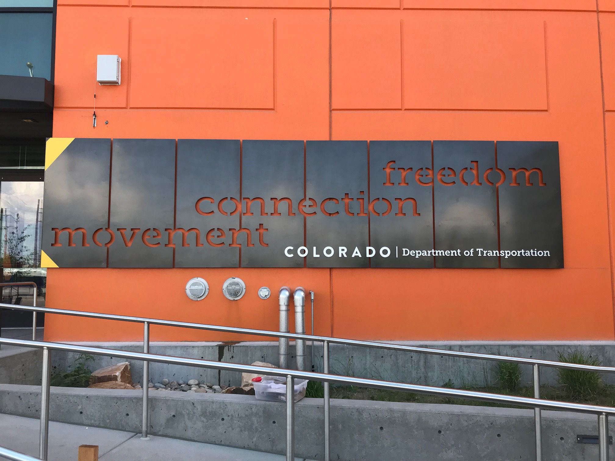 A sign on an orange wall reads "movement, connection, freedom" with cut-out letters. Below, it reads "Colorado Department of Transportation" in white.
