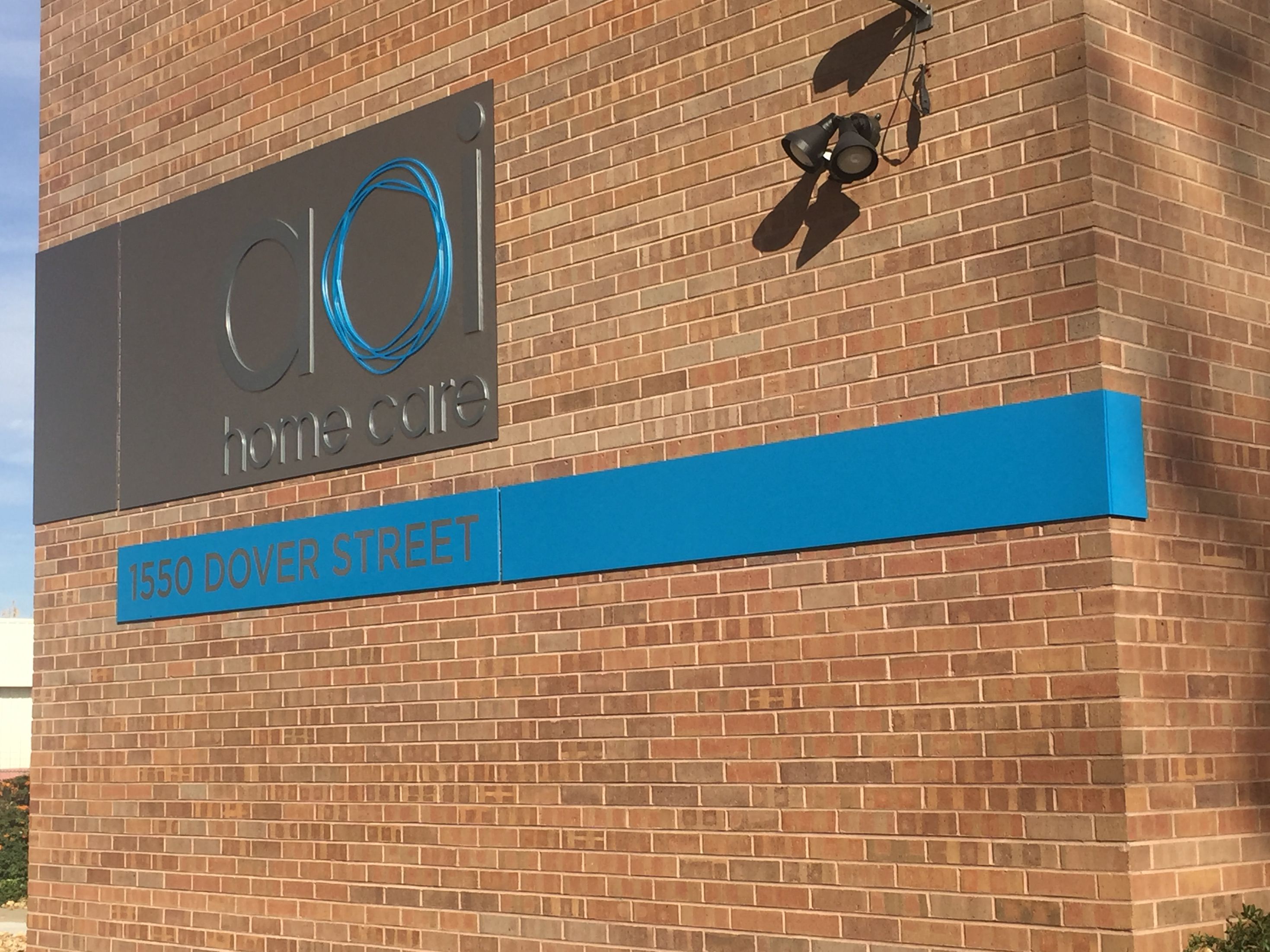 A brick building wall displaying a sign for "AOI Home Care" with a logo of two blue circular shapes. Below the sign, a blue banner reads "1550 Dover Street." A black security camera is mounted above the sign.