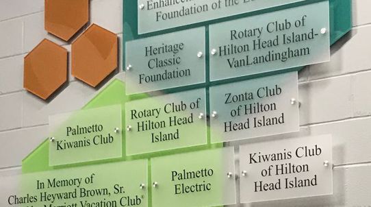 A wall display with various plaques recognizing different organizations and individuals. The plaques include names like "Rotary Club of Hilton Head Island," "Palmetto Electric," and "Kiwanis Club of Hilton Head Island," among others. Three orange hexagon shapes are mounted on the top left.