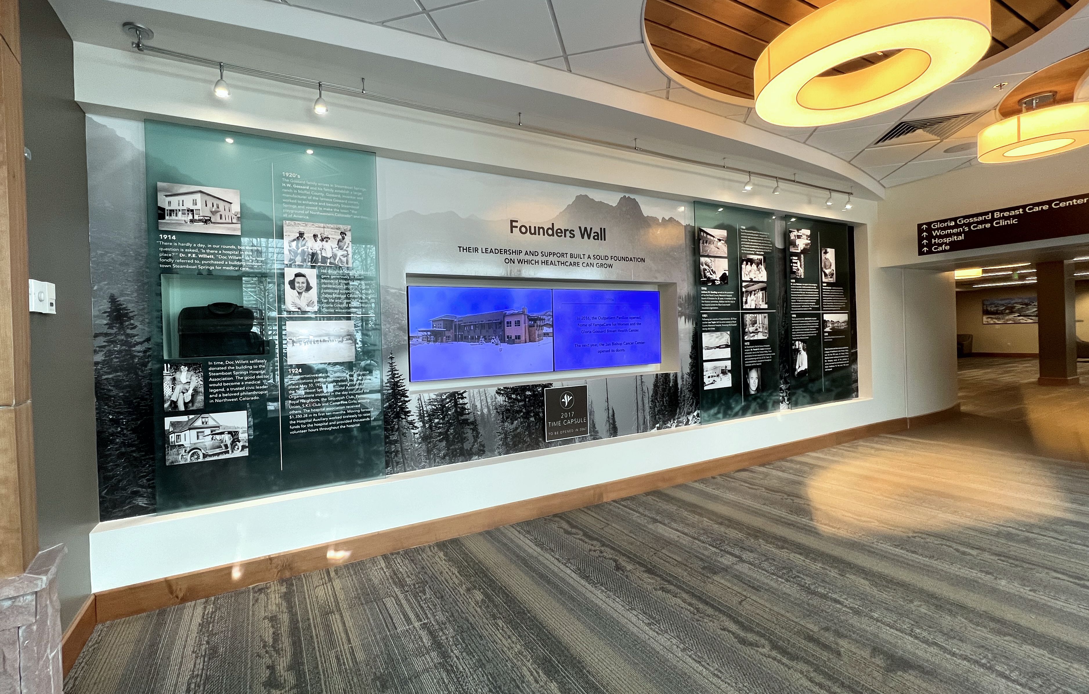 A well-lit hallway features a "Founders Wall" display with historical photos, text, and a blue-tinted screen. The wall honors influential figures in the establishment of the institution. Modern light fixtures hang from the ceiling and signs direct to various rooms.