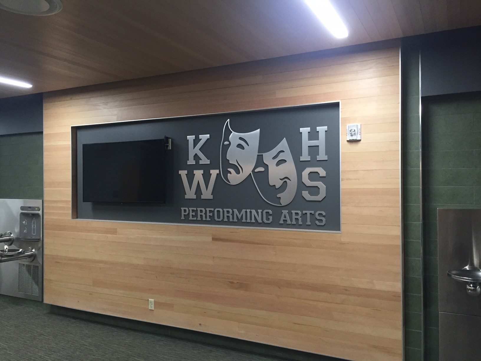 A dark wall with silver metal letters "KWHS Performing Arts" arranged around two theatre masks—one smiling and one frowning—symbolizing comedy and tragedy. The wall is framed by light wood paneling.