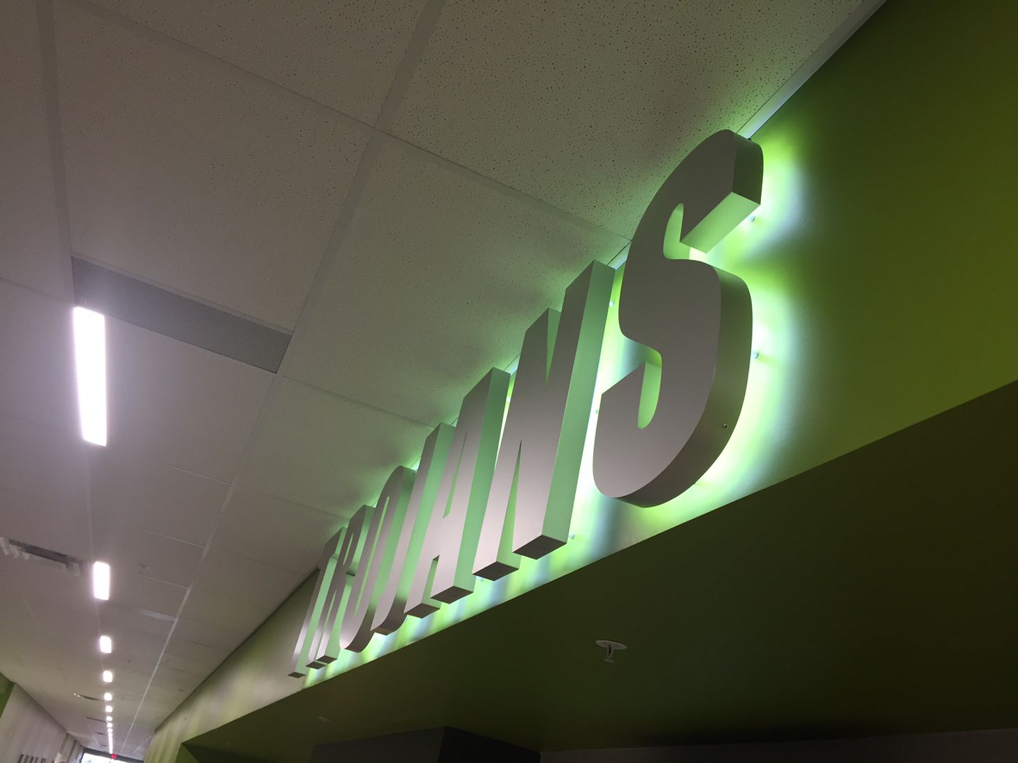 A hallway ceiling with overhead lighting and a large 3D sign reading "TROJANS" mounted on a green wall, illuminated with a green backlight. The corridor has a modern, clean appearance with minimalistic design elements.