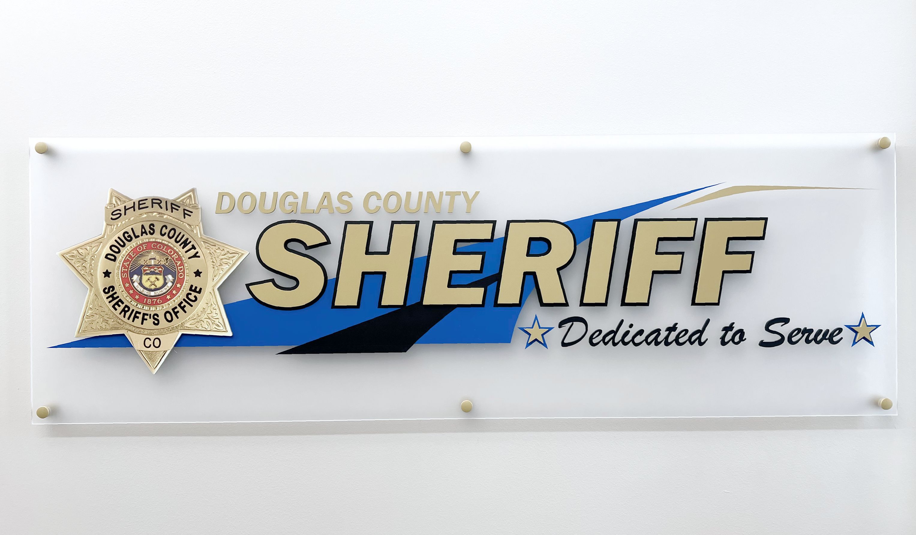 A large sign on a wall reads "Douglas County Sheriff Dedicated to Serve." The text is accompanied by a sheriff's badge on the left side, and the background features black and blue accents.