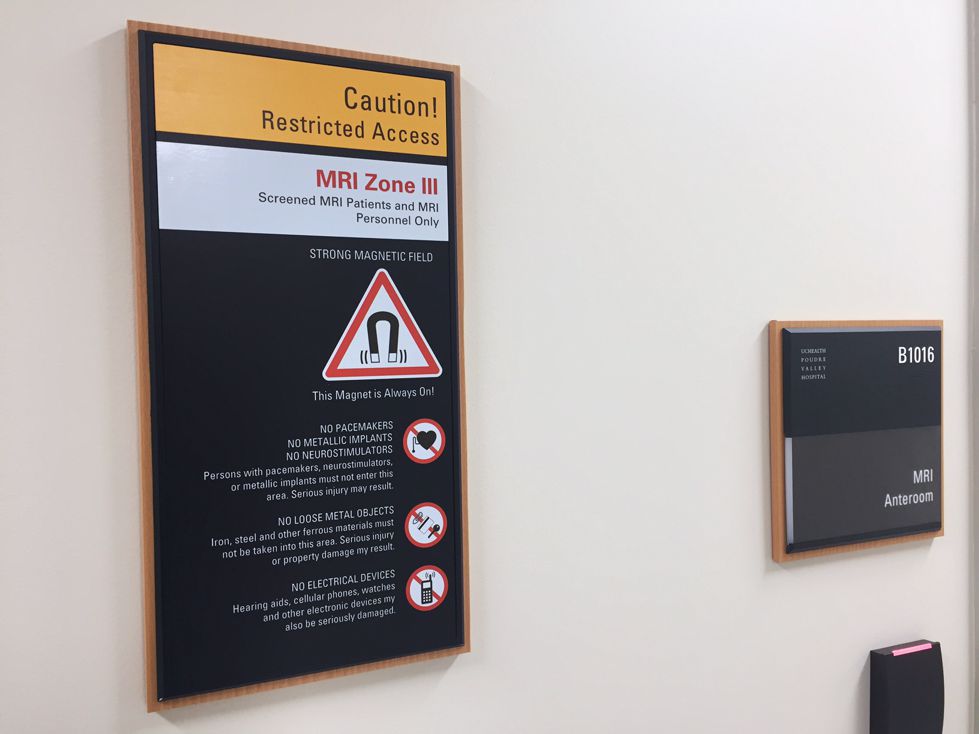Two informational signs in English and Spanish are displayed on a wall. They provide guidelines and warnings for entering MRI Zone II. Each sign has a blue and black background with white text and includes magnetic field hazard symbols.