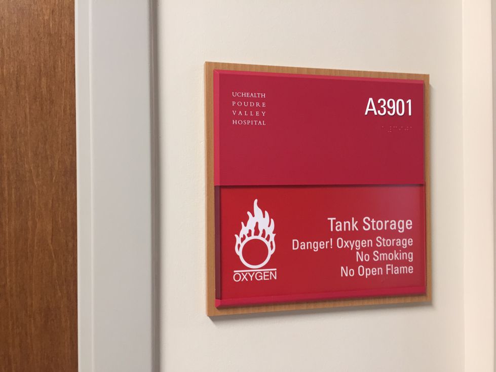 A red sign on a wall outside a room labeled "A3901" at UCHealth Poudre Valley Hospital. The sign reads: “Tank Storage. Danger! Oxygen Storage. No Smoking. No Open Flame,” accompanied by a warning symbol of flames above the word "OXYGEN.