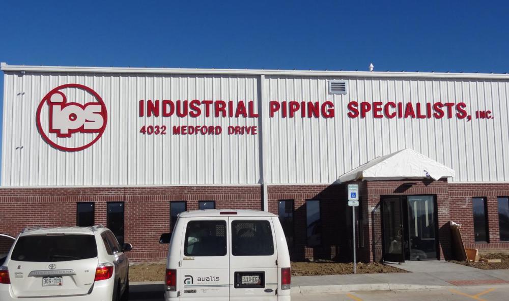 A large industrial building with "IPS" logo and text reading "Industrial Piping Specialist, Inc." on the white upper facade above red brick lower walls. The building is located at "4025 Menoah Road" with blue sky in the background.