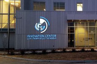 The image shows the exterior of the Innovation Center for St. Vrain Valley Schools. The building has metal siding with large windows. A large illuminated sign featuring the center's name and logo is prominently displayed on the building's wall.