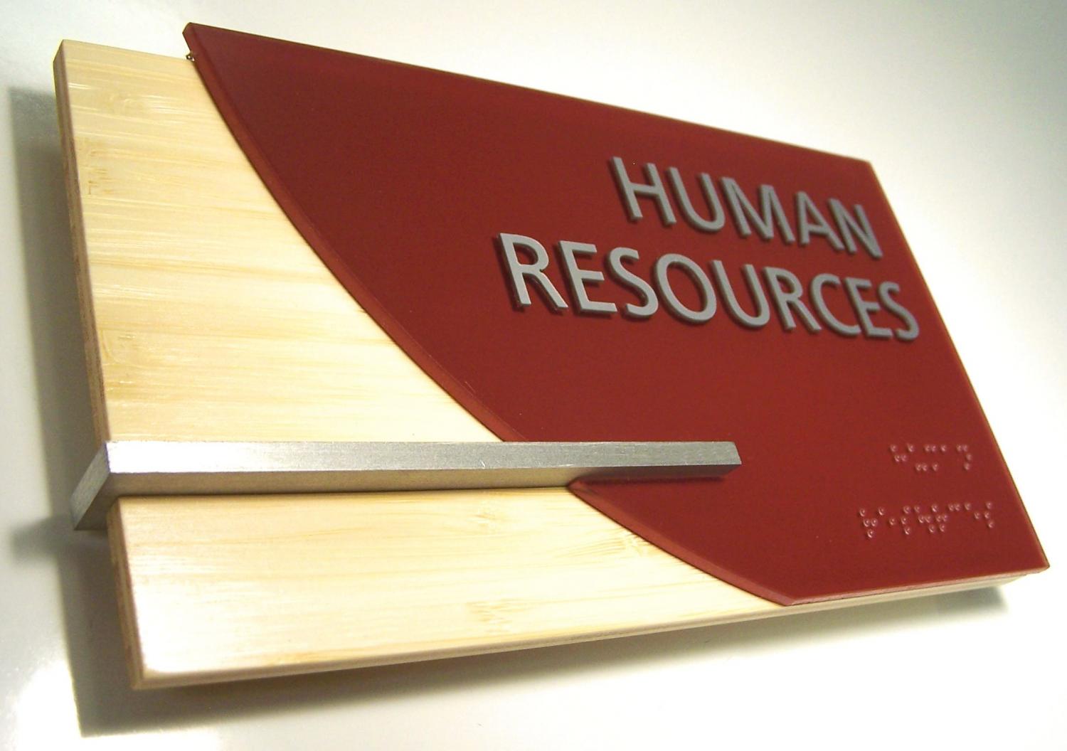 A wall-mounted sign with "Human Resources" written in large white letters on a red background. The sign also includes Braille text at the bottom. The sign has a wooden part on the left side with a horizontal metal bar extending halfway across.