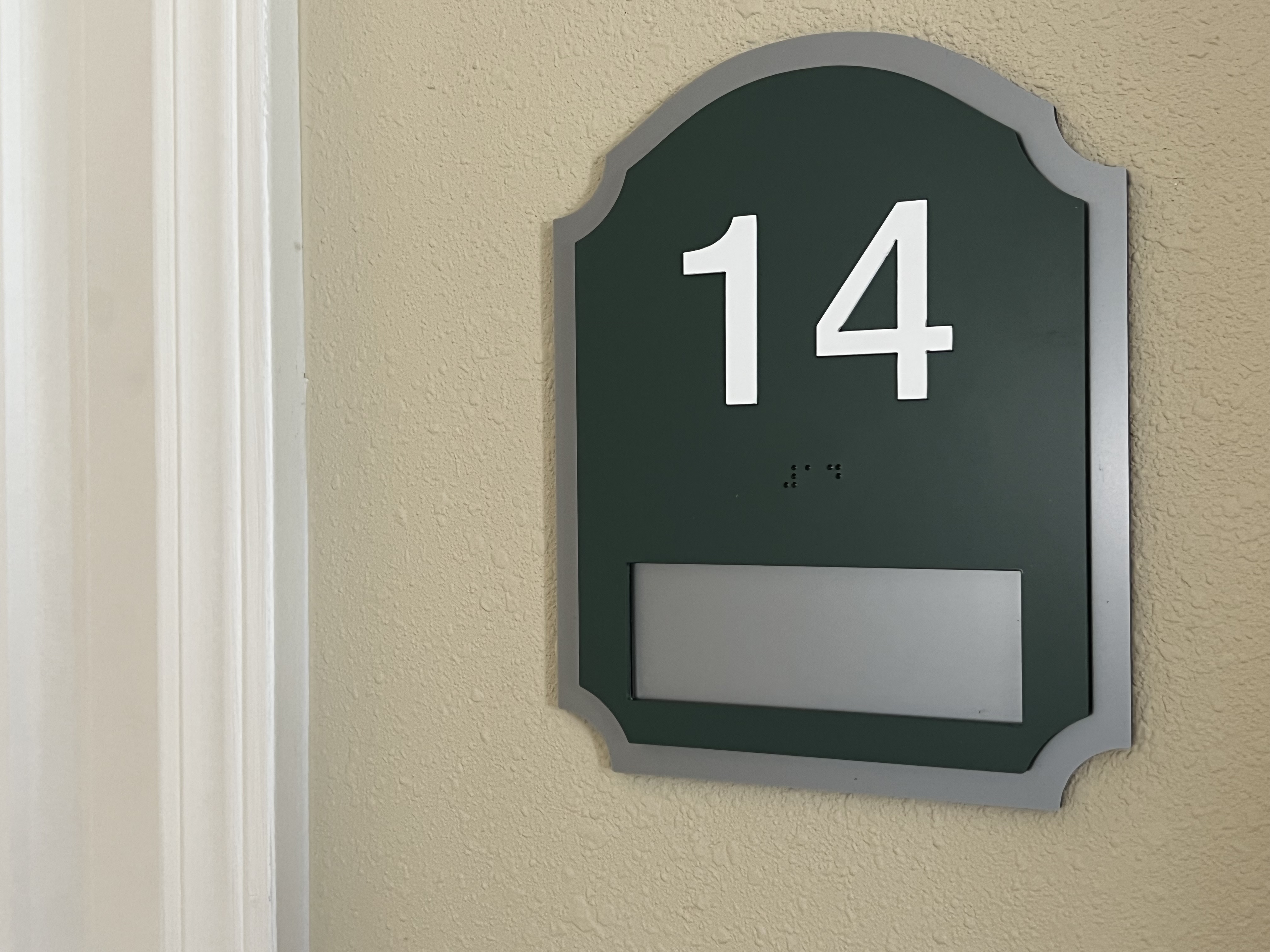 A green and gray door sign with the number 14 prominently displayed in white. Below the number, there is a small line of braille dots. There is a blank rectangular slot near the bottom of the sign.