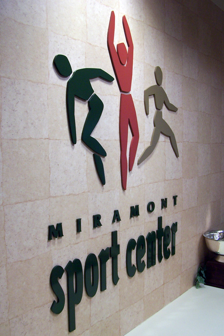 Wall sign for Miramont Sport Center featuring three stylized human figures in green, red, and brown engaged in sports activities. The words 'Miramont sport center' are prominently displayed beneath the figures.  This is a close-up view of the sign.
