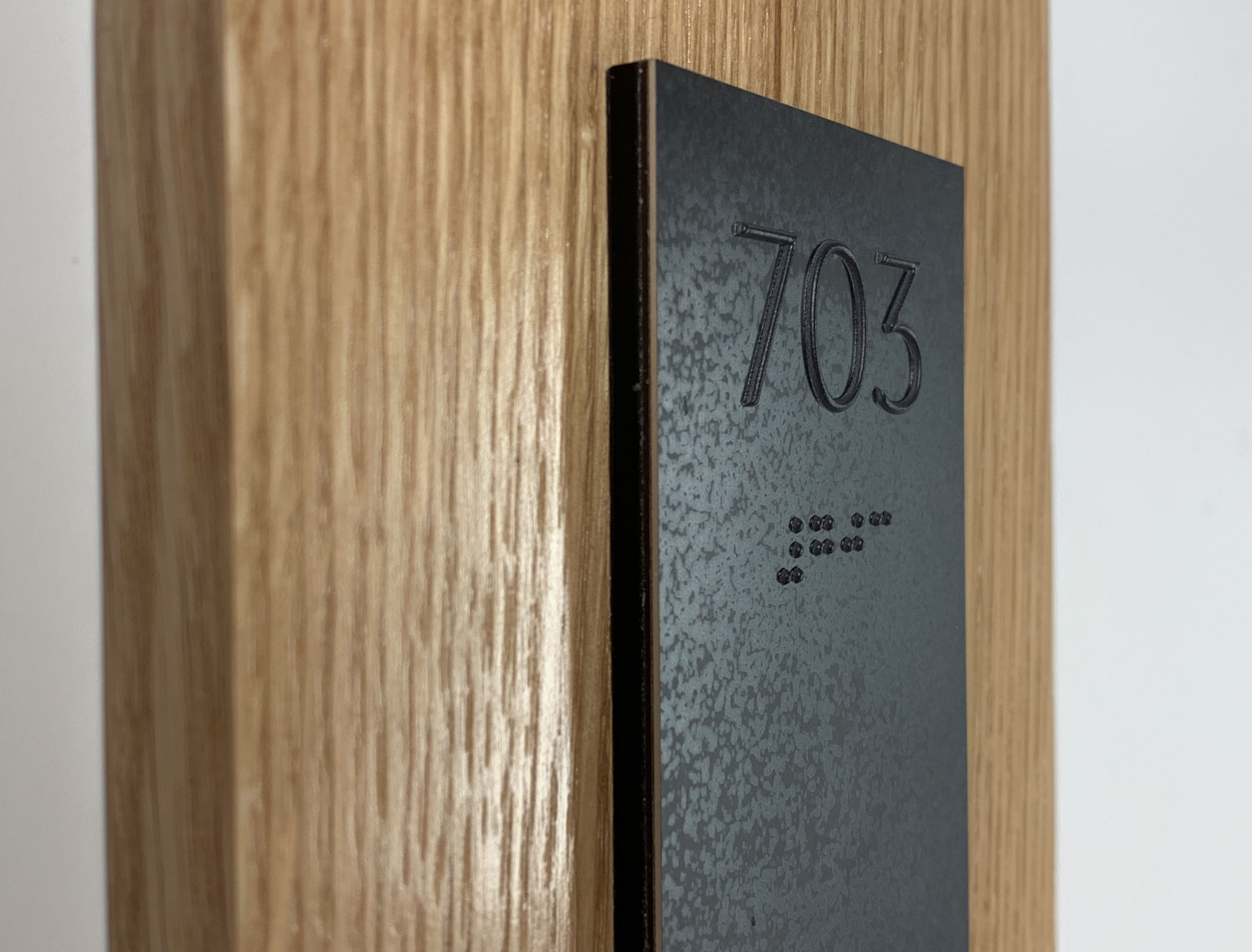 A dark tactile sign with the number "703" and Braille characters below it is mounted on a wooden surface. The image is taken from a side angle, showing the texture and depth of the sign.