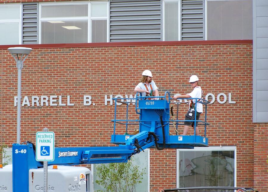 Two workers in hard hats use a blue cherry picker to install or repair signage on a brick building labeled "HARRELL B. HOWE SCHOOL". The scene is outdoors and includes a reserved parking sign in the foreground.
