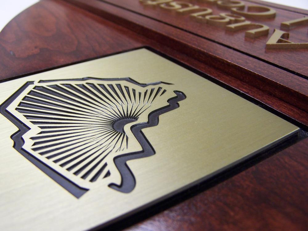 Close-up of a wooden plaque with a gold-colored metal plate on it. The plate features an engraved abstract design resembling the outline of a state with intricate lines radiating from its center. Part of an engraved inscription is visible along the top edge.