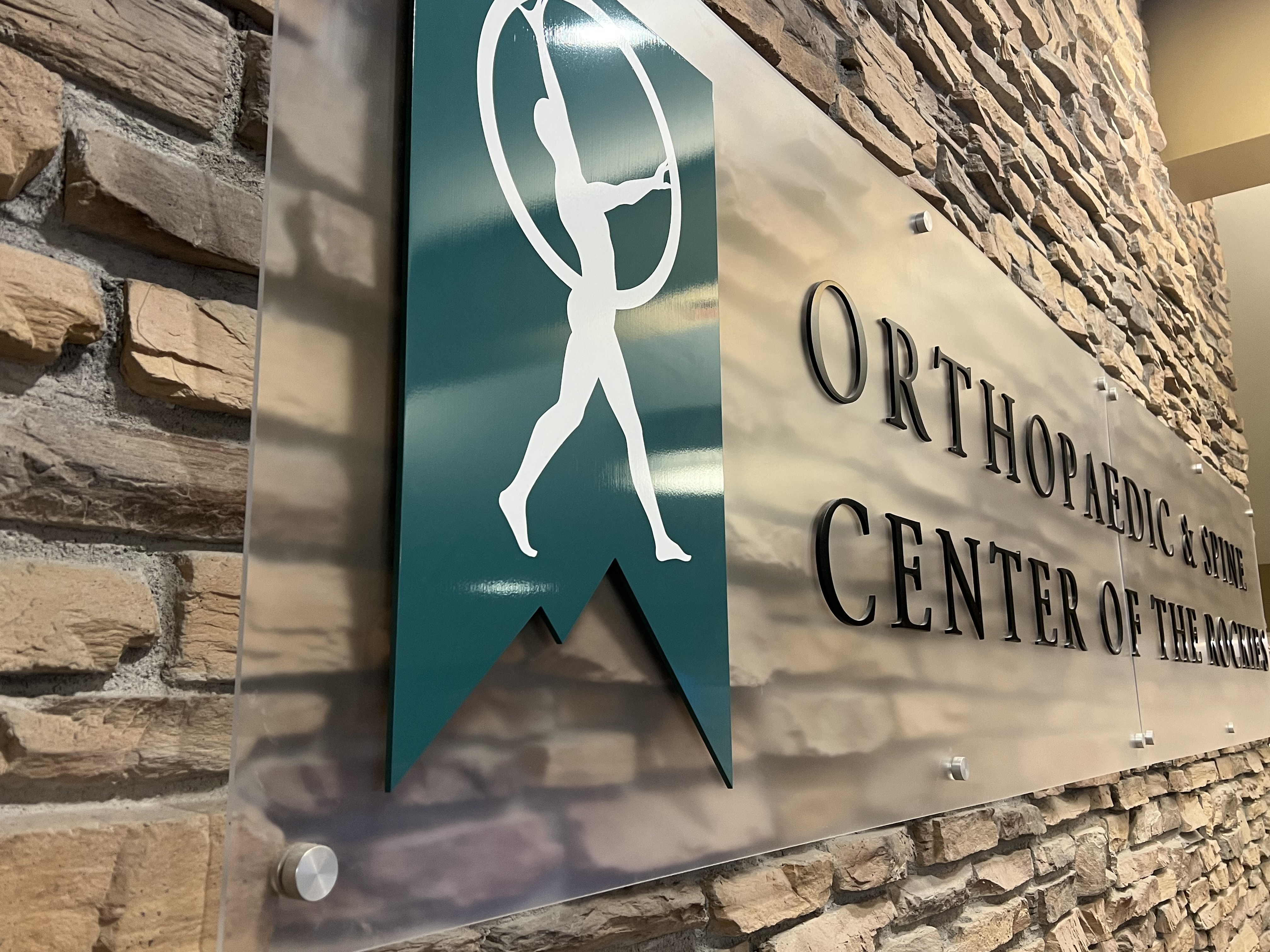 A sign on a stone wall reads "Orthopaedic & Spine Center of the Rockies." The sign features a graphic of a person  within a circular frame. Multiple screws affix the clear sign panel to the wall.