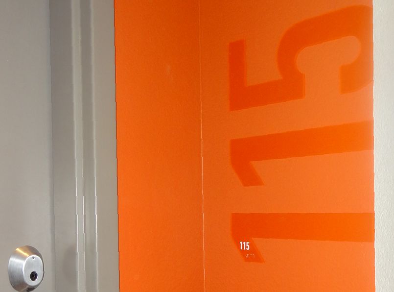 A close-up of a room door with bright orange paint and large numbers "115" displayed prominently. The door handle, metallic and round, is visible on the left side of the image.