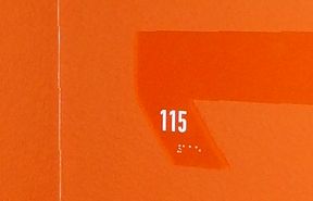 An orange wall with the number "115" written in white on a flat, slightly raised rectangular surface. Beneath the number, there are small braille dots for tactile reading. The image captures a corner section of the wall.