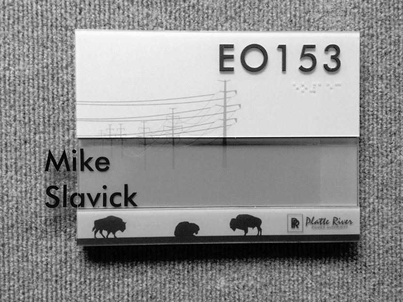 Office nameplate on a gray fabric background, featuring the text "EO153" and "Mike Slavick." The design includes illustrations of power lines and three buffalo silhouettes at the bottom. The Platte River logo is also visible.