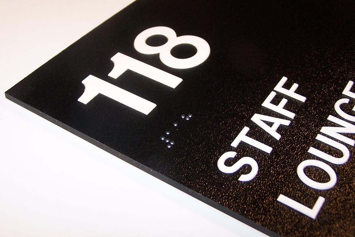 Close-up of a black and white room sign. The number "118" is prominently displayed at the top in large white font. Below the number in smaller white text are the words "STAFF LOUNGE." Braille dots are positioned between the number and the text.