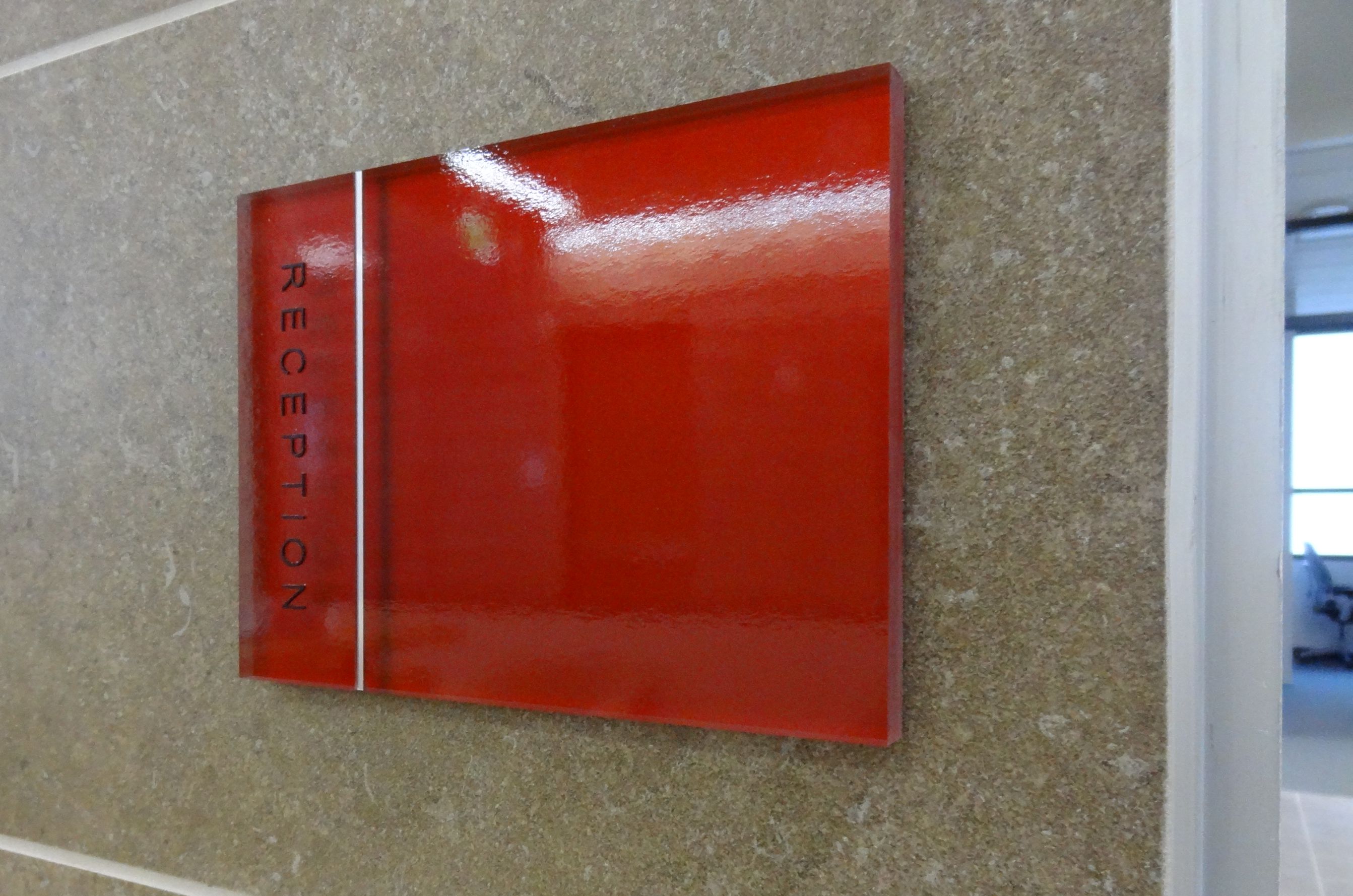A red sign mounted on a beige, tiled wall with the word "RECEPTION" written vertically in black letters on the left side. The sign has a glossy finish and is situated next to a hallway opening with visible natural light.