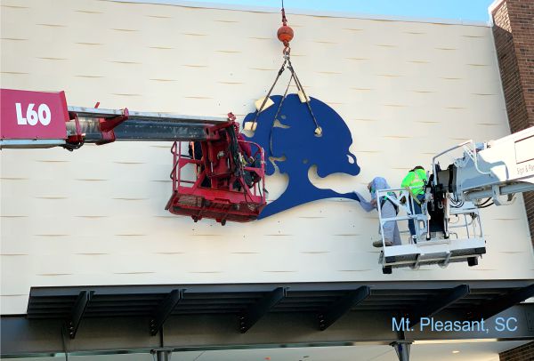 A blue metal silhouette of a tree logo is being installed on a building wall in Mt. Pleasant, SC. Workers in two lift baskets are securing the sculpture with the help of a crane. The background shows the cream-colored exterior of the building.
