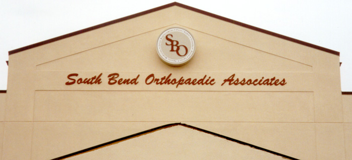 The image shows the exterior of a building with a beige facade. At the top, there's a circular logo with the letters "SBO." Below the logo, the building is labeled "South Bend Orthopaedic Associates" in red cursive text.