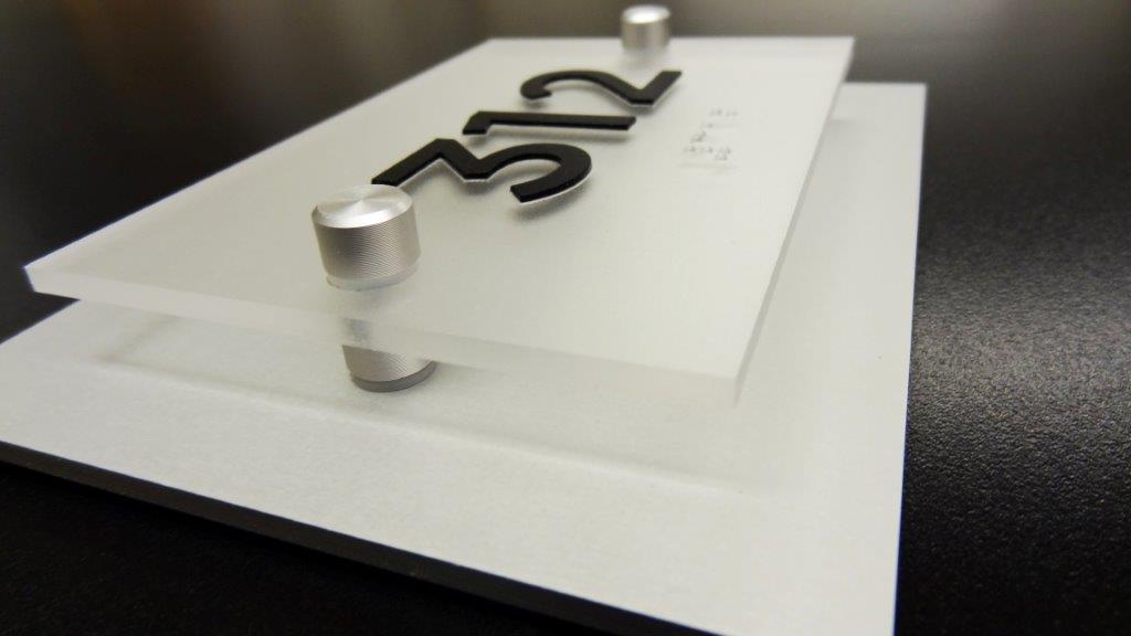 Close-up of a modern office door sign displaying the number 312. The sign consists of layered frosted acrylic panels with black raised numbers on the top panel. The panels are slightly offset and held together by metallic cylindrical standoffs.