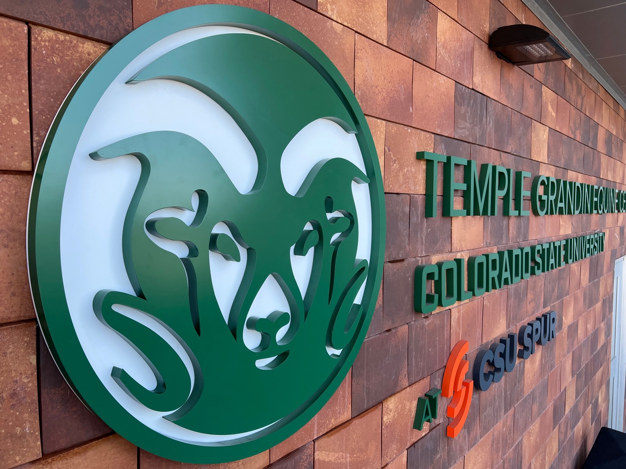 Exterior wall of the Temple Grandin Equine Center at Colorado State University. The wall features the CSU Rams logo and the facility's name in green text, along with the CSU Spur logo.