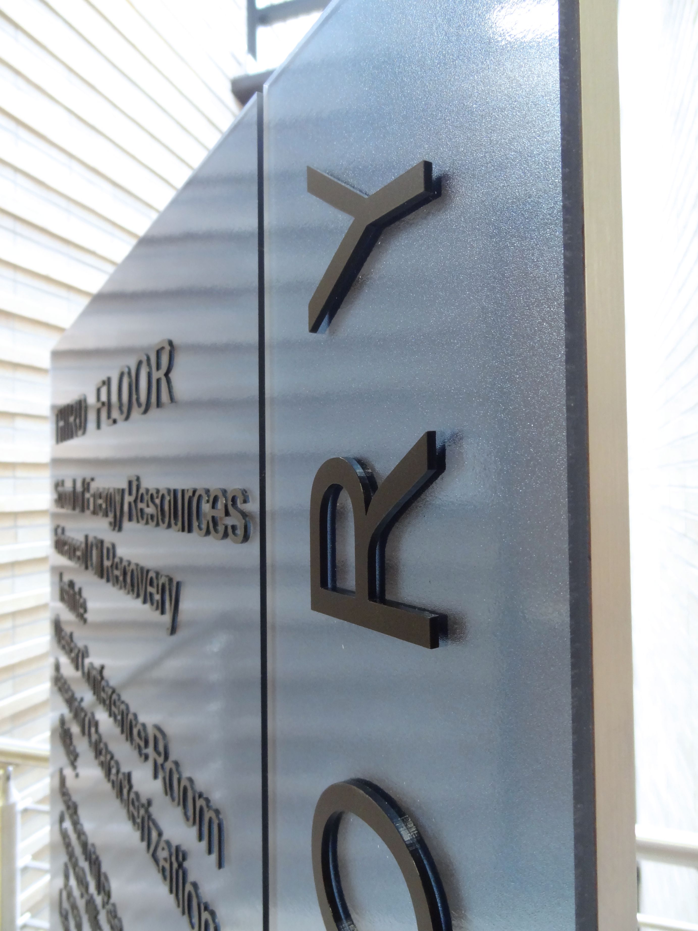 Close-up of a large, vertical directory sign in a modern building. The sign is made of sleek metal with raised, glossy black text listing various facilities such as library resources and financial recovery. The background is blurred, revealing part of the building’s interior.