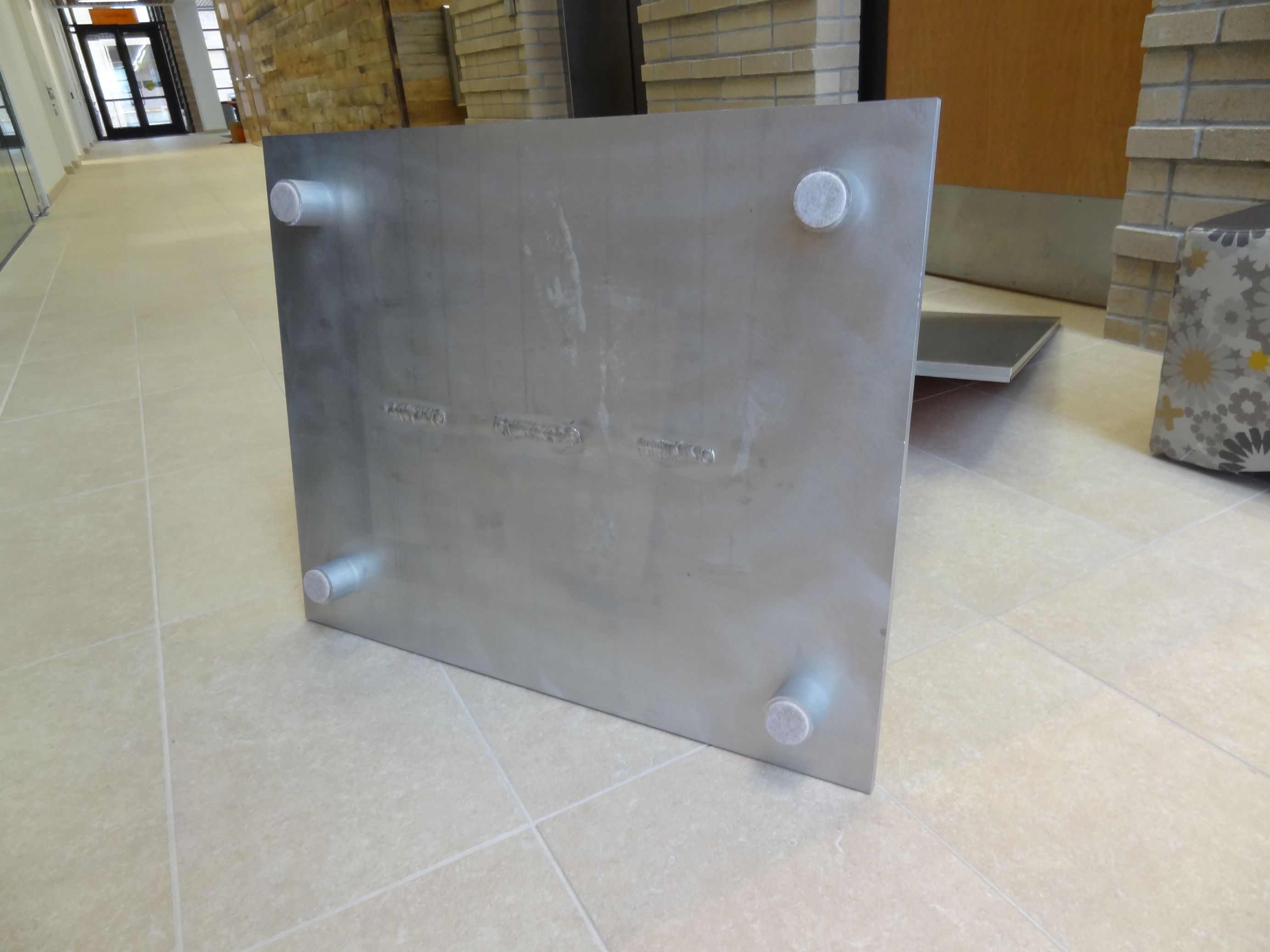 A metal plate with four cylindrical feet is positioned upright on a tile floor in a building hallway, showing the underside of a floor standing directional sign.