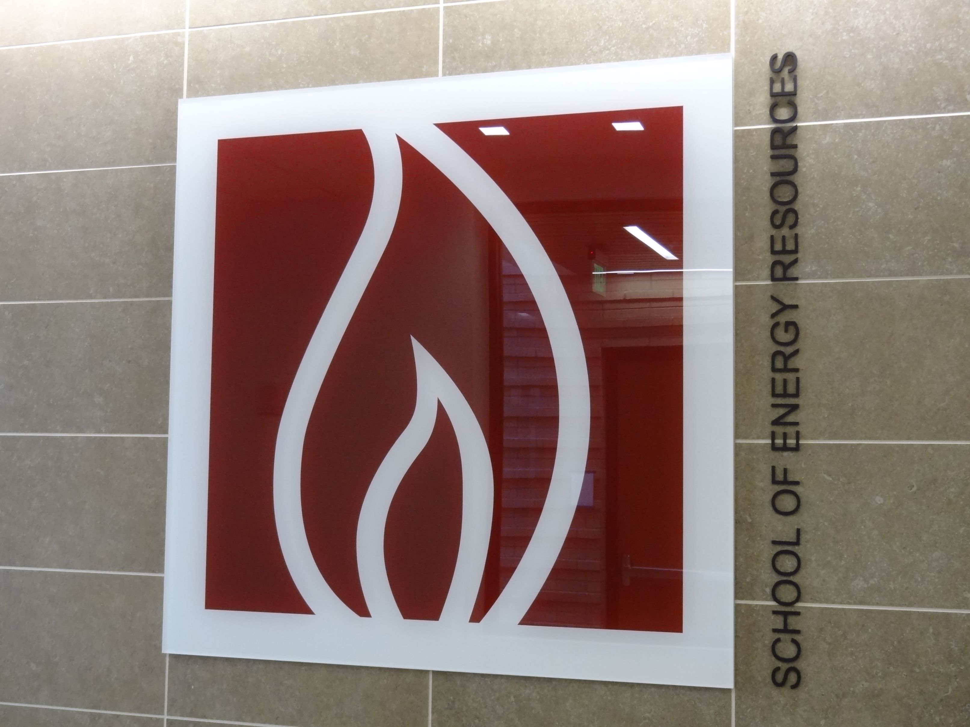 A sign on a wall for the School of Energy Resources. The sign features a stylized flame in white against a red background. The words "School of Energy Resources" are written vertically along the right side of the sign. The wall is tiled in a neutral color.