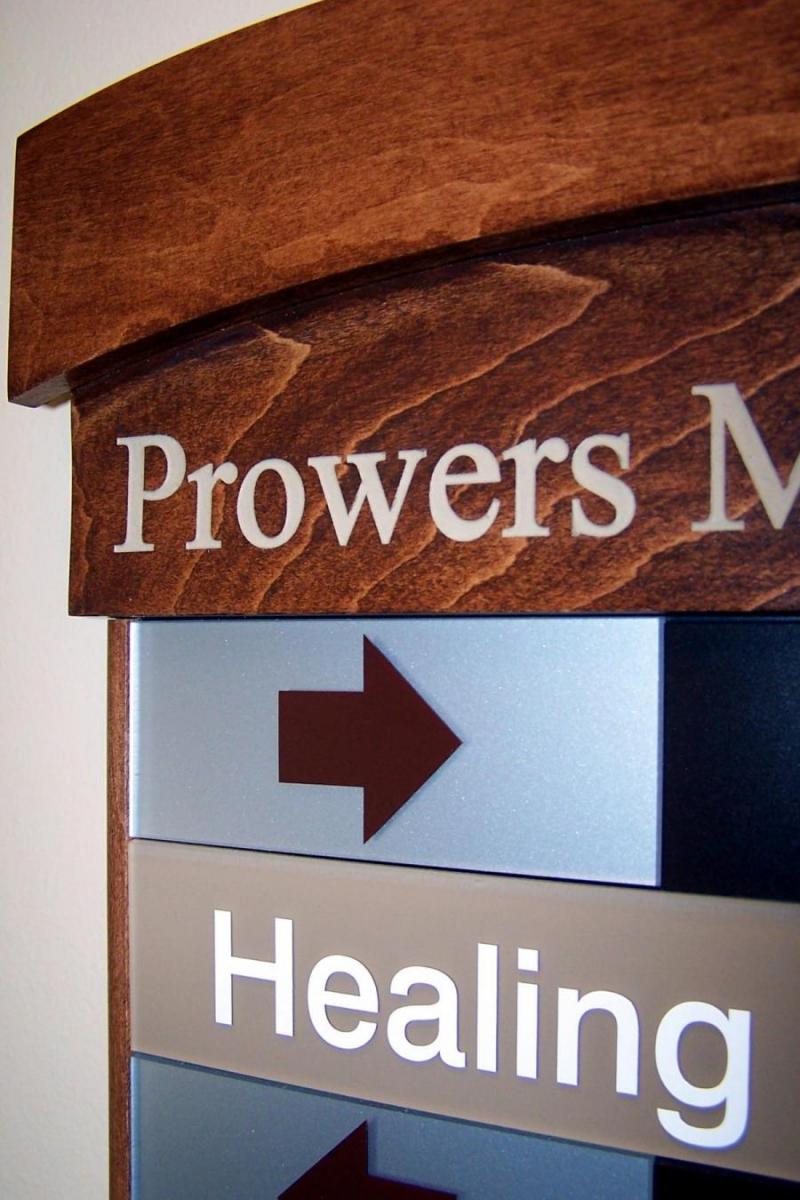 A close-up view of a directional sign with a wooden frame. The top part of the sign partially shows the words "Prowers M" and below it features a large arrow pointing left, followed by the word "Healing" on a gray background.