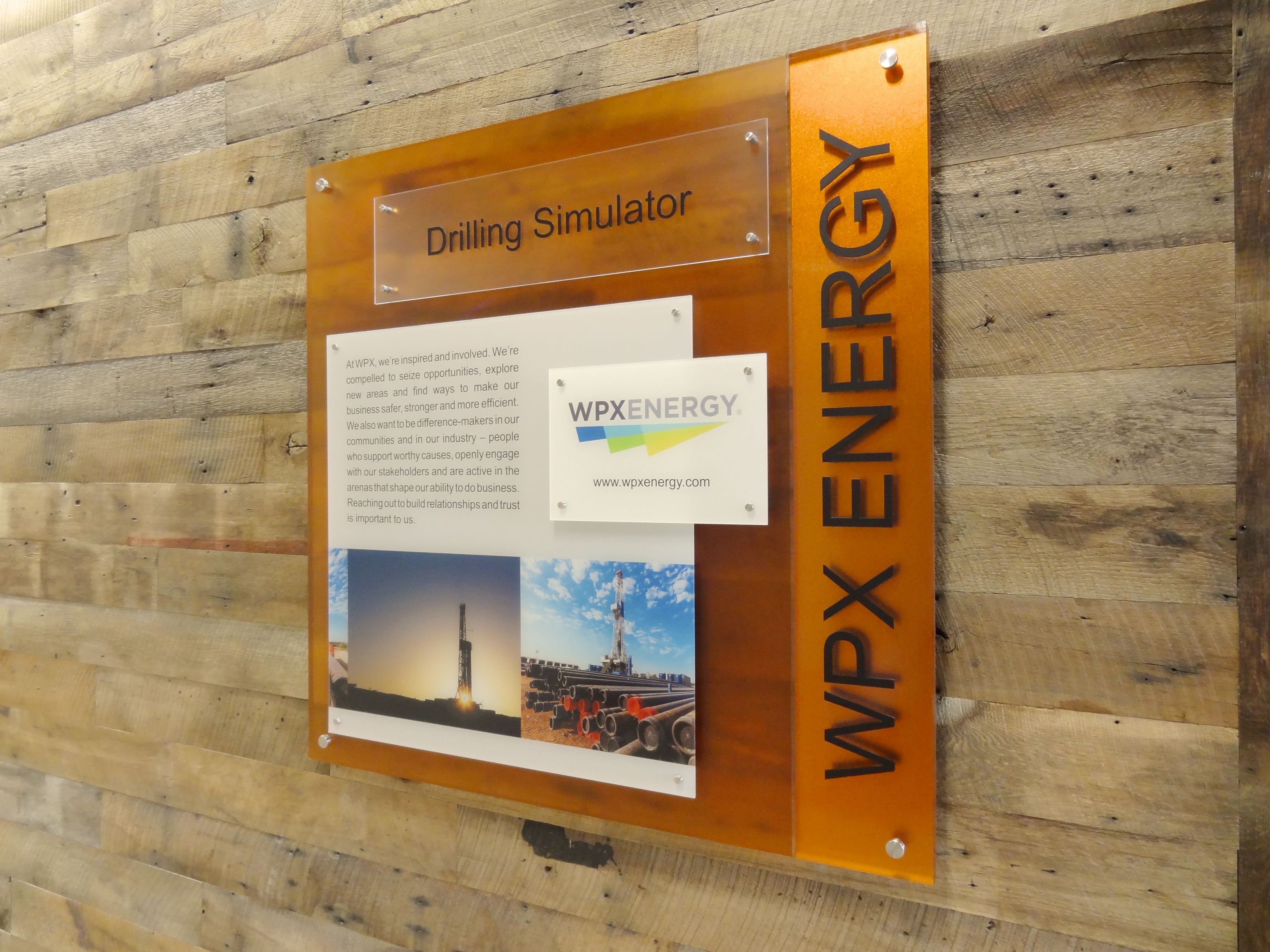 A wooden wall with a sign reading "Drilling Simulator" and "WPX ENERGY." The sign includes images of oil rigs and text about the company's work. The WPX ENERGY logo is visible, featuring a blue and green swoosh design.