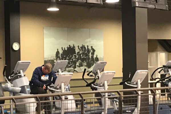 A gym with a row of exercise bikes are lined up next to a divider. A large black-and-white photograph of a group of people is mounted on the beige wall in the background.