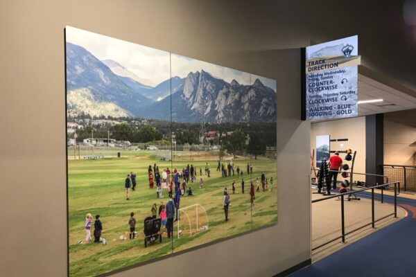 A hallway with a large photograph on the wall depicting people playing sports on a field with mountains in the background. To the right, exercise equipment and individuals working out can be seen in an adjacent room. A sign hangs from the ceiling with directions.
