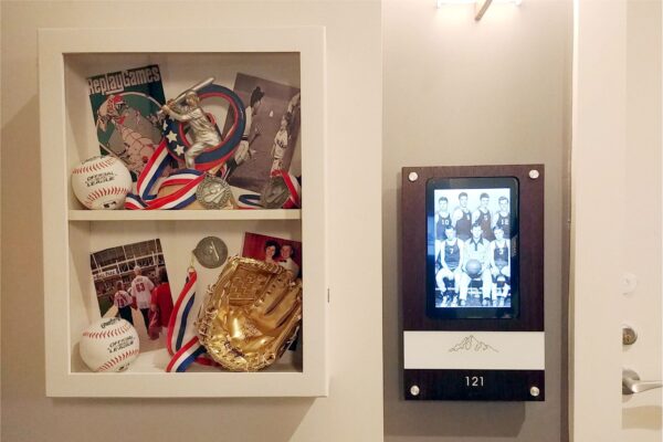 A display case with sports memorabilia, including baseballs, a baseball glove, medals, and sports photographs is shown on the left. On the right, there is a digital picture frame displaying a team photo and the number "121" below it.