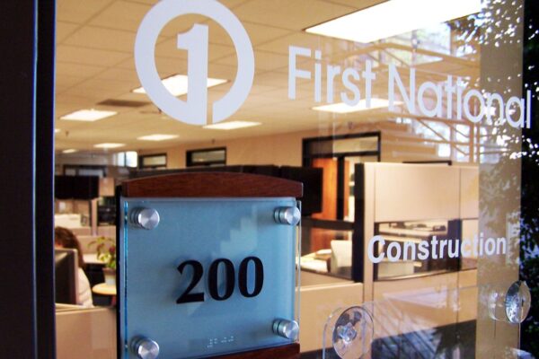 Office entrance with a glass door bearing the sign "First National Construction" and a frosted glass plate with the number "200" on it. Inside, there are cubicles and visible office equipment.