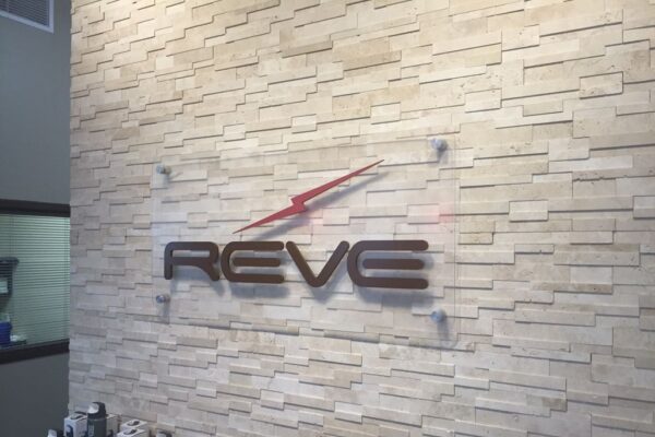 A wall with a textured stone finish featuring a clear sign that reads "REVE" with a red arrow-like design above the text. Below the sign, several bottles and products are displayed on a shelf.
