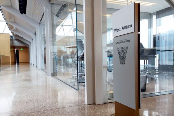 A modern hallway with floor-to-ceiling glass walls on the right, leading to a sunlit atrium labeled "West Atrium" on a sign. Inside the glass-walled room are high chairs and a table, with a polished concrete floor extending through the area.