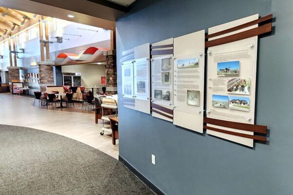 A display on a dark-colored wall features four acrylic panels with historical text, images, and dates ranging from 1869 to 1920. Each panel is bordered by wooden slats, and the first panel caption reads, "We all begin somewhere. Here is our story.