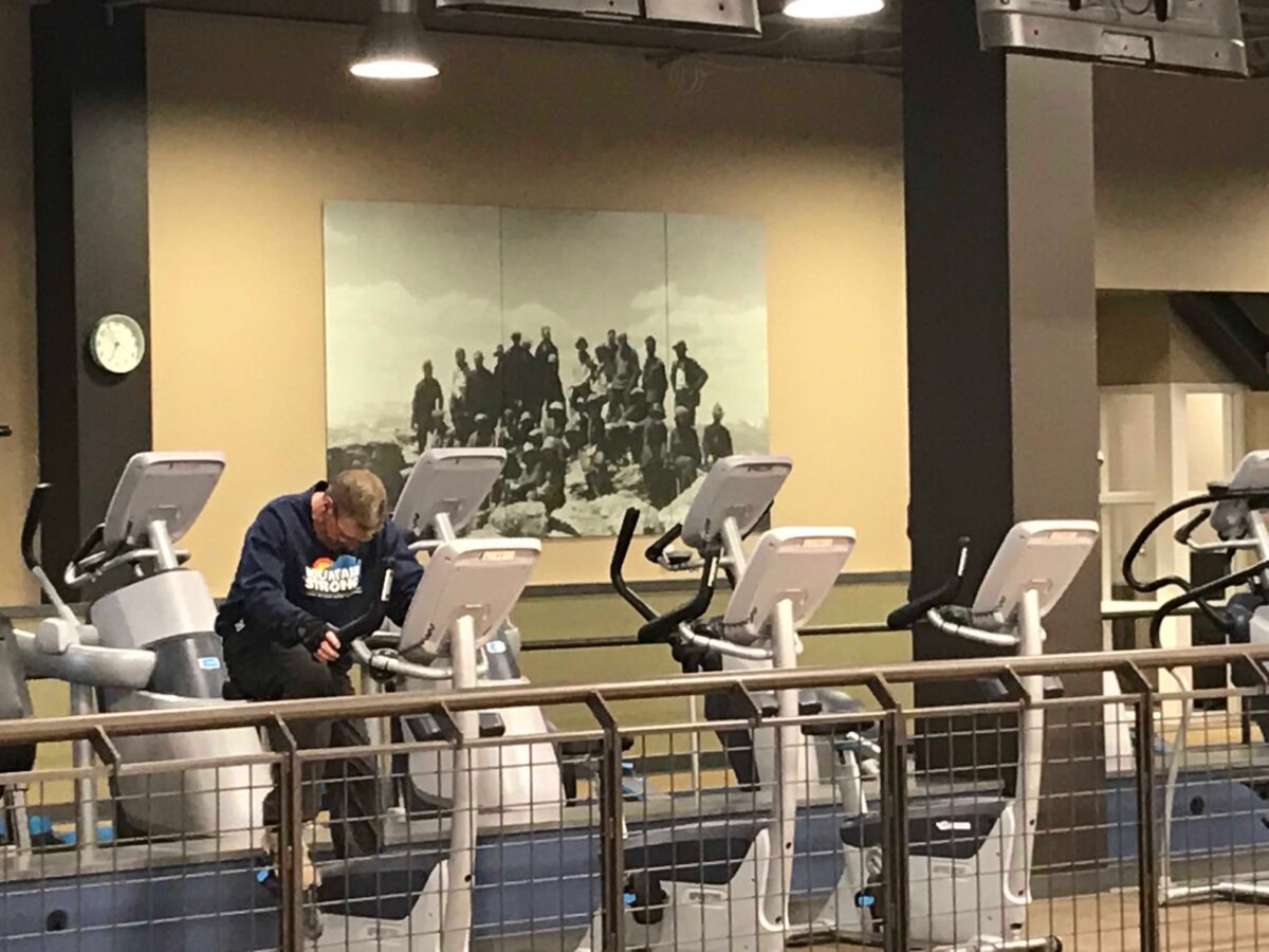 A gym with a row of exercise bikes are lined up next to a divider. A large black-and-white photograph of a group of people is mounted on the beige wall in the background.