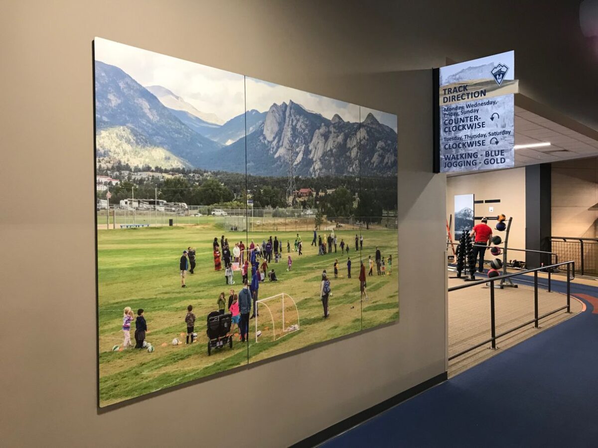 A hallway with a large photograph on the wall depicting people playing sports on a field with mountains in the background. To the right, exercise equipment and individuals working out can be seen in an adjacent room. A sign hangs from the ceiling with directions.