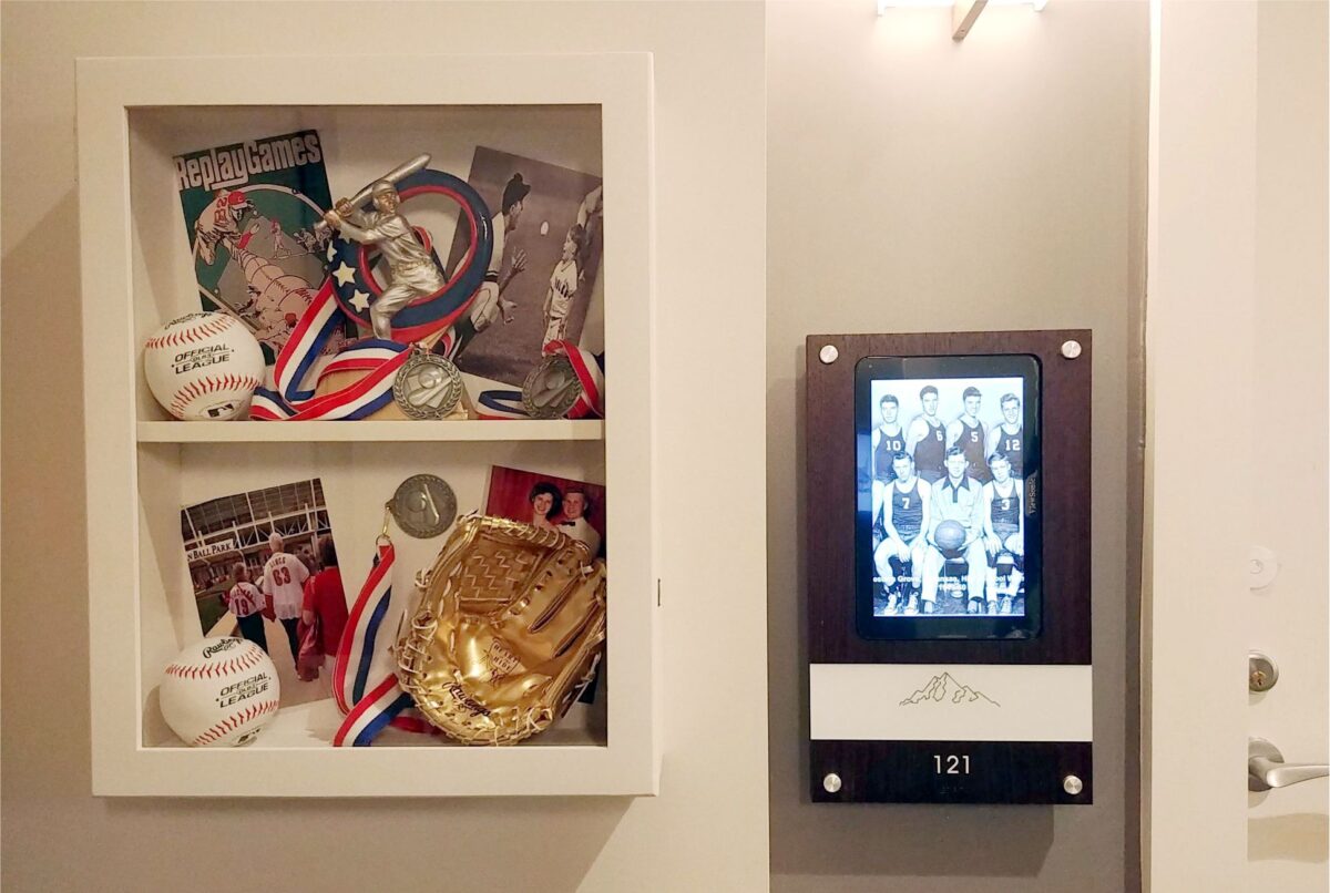 A display case with sports memorabilia, including baseballs, a baseball glove, medals, and sports photographs is shown on the left. On the right, there is a digital picture frame displaying a team photo and the number "121" below it.
