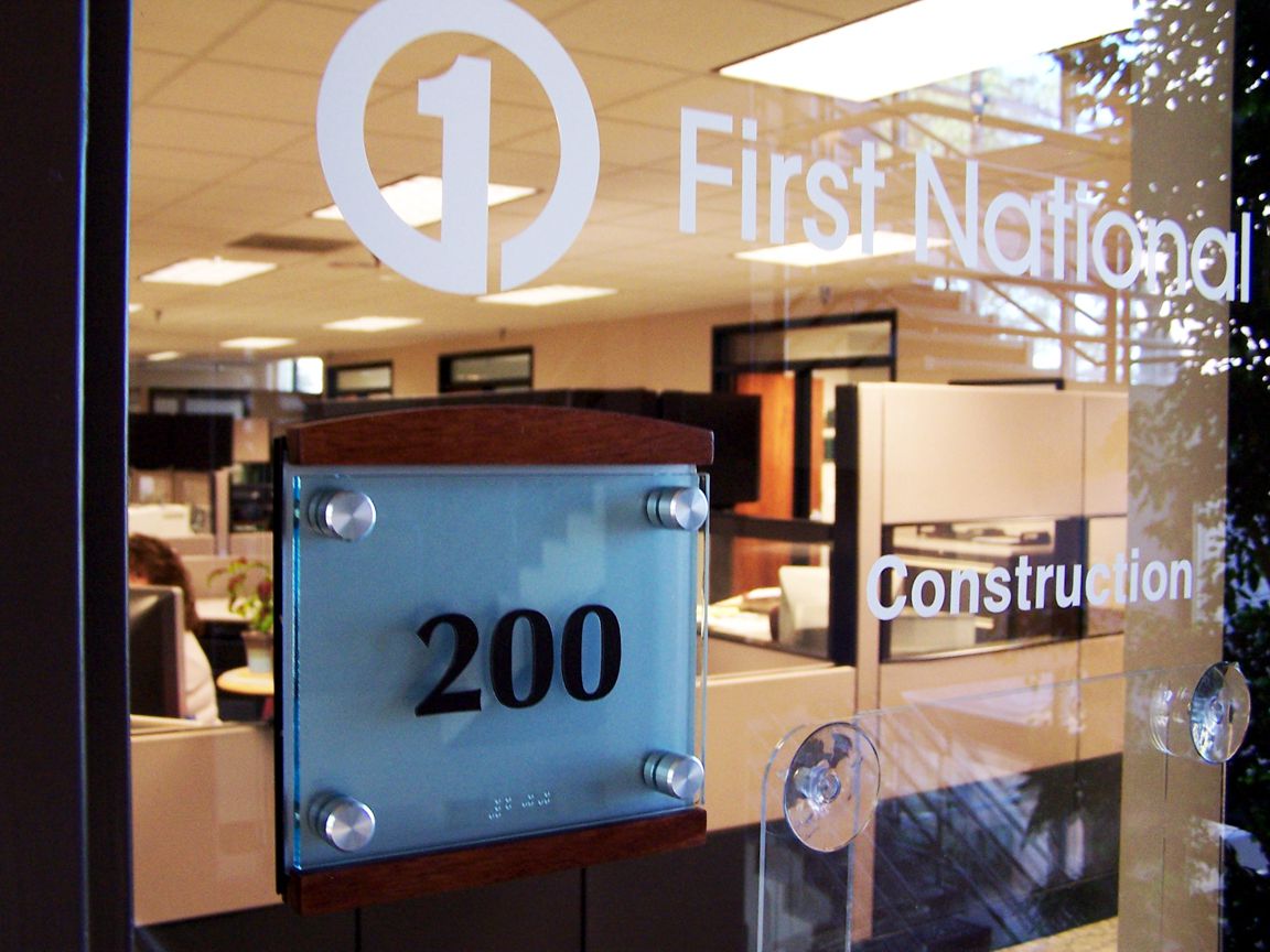 Office entrance with a glass door bearing the sign "First National Construction" and a frosted glass plate with the number "200" on it. Inside, there are cubicles and visible office equipment.
