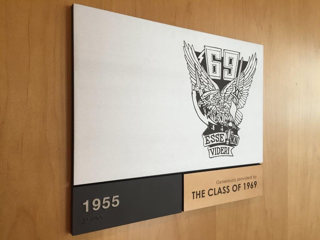 A plaque mounted on a wooden wall features a logo with an eagle, the number "69", and the words "Essel LG Viden." Below the image, a metallic strip reads "1955" and "Presented by the class of 1969.
