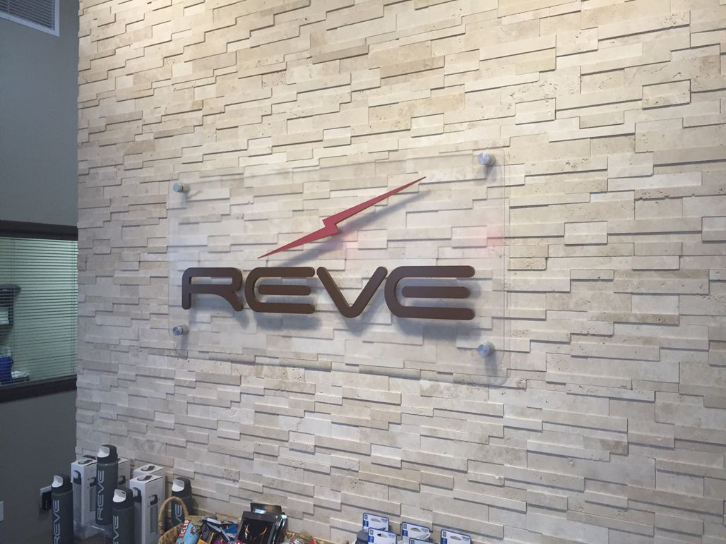 A wall with a textured stone finish featuring a clear sign that reads "REVE" with a red arrow-like design above the text. Below the sign, several bottles and products are displayed on a shelf.
