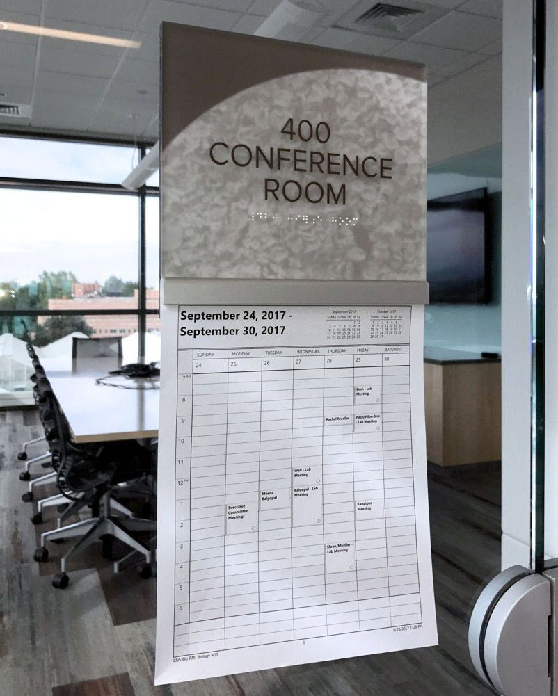 A wall sign reads "400 Conference Room" in text and Braille. Below it, a weekly schedule for September 24 - 30, 2017, is posted, detailing daily meetings and events. The conference room contains a long table with chairs and a large screen in the background.
