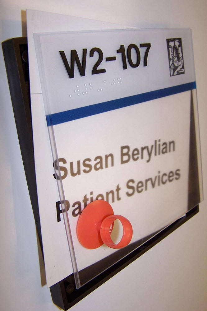 A room sign with the text "W2-107" and "Susan Berylian Client Services" is mounted on a wall. The sign has a blue stripe and a small icon in the top right corner.  Braille text is above the blue stripe.