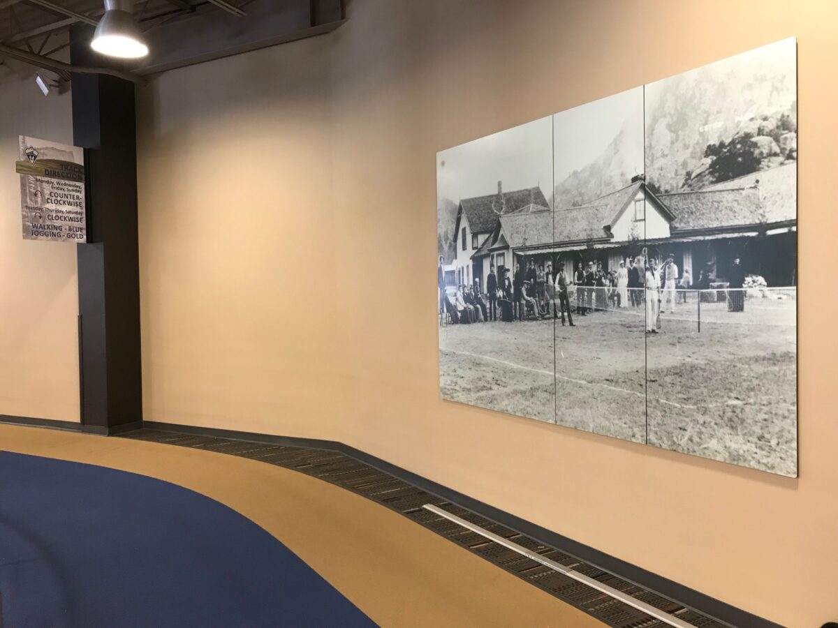 A large black-and-white historical photograph hangs on a light beige wall in a spacious room with high ceilings. The floor has a curving path design with a combination of blue and tan tiles. The photo depicts a group of people standing outside old buildings.