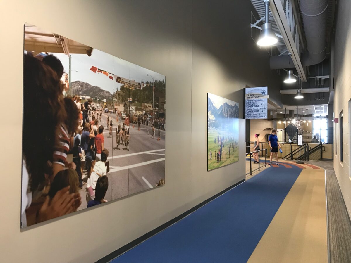 A spacious indoor hallway features large photos on the walls. On the left is an image of people observing an outdoor event. The hallway has overhead lights and a blue and beige walkway.